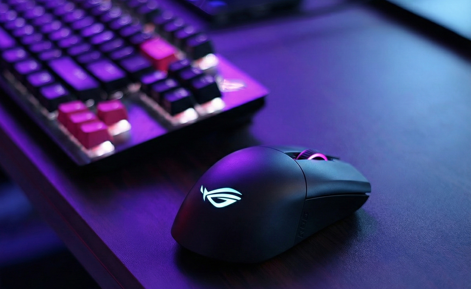Ratón gaming ASUS ROG Keris Wireless color negro en un escritorio gamer iluminado con luces neón verdes y moradas, junto a un teclado mecánico y monitor.
